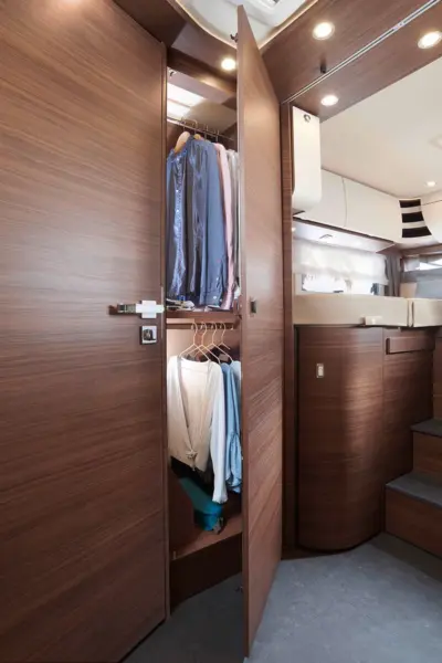 Ein moderner Holzschrank mit offenen Türen offenbart ordentlich aufgehängte Hemden und Blusen in einem kompakten, stilvollen Raum der Carthago Chic C Line mit geschwungenen Wänden und Stufen, möglicherweise in einer Jacht oder einem Wohnmobil.