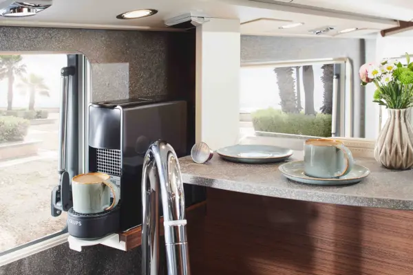 Moderne Küche in einem Carthago Chic C Line T mit einer Kaffeemaschine, die zwei Tassen auf dem Tresen in der Nähe eines Fensters zubereitet; zwei grüne Tassen und Teller, eine Blumenvase und ein Blick ins Freie mit Palmen sind zu sehen.