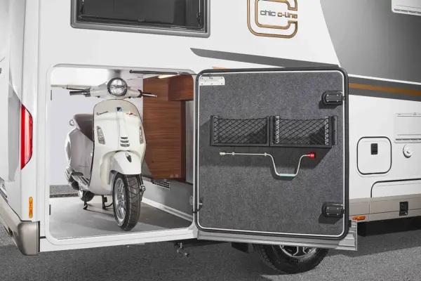 Ein weißer Roller wird im Staufach an der Rückseite eines Carthago Chic C Line Wohnmobils geparkt, wobei die Tür des Fachs geöffnet ist, um einen geordneten Stauraum und den Zugang zum Roller zu ermöglichen.