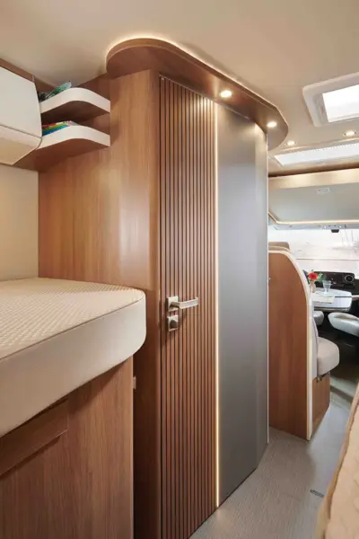 Modernes Wohnmobil-Interieur mit Holzvertäfelung, eleganter Schiebetür, eingebautem Dachboden, gepolstertem Bett auf der linken Seite und Blick auf die Fahrerkabine mit Oberlicht und Sitzgruppe - charakteristische Merkmale des Carthago C2-Tourer.