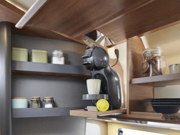 Eine carthago c2-tourer Kaffeemaschine steht auf einem Holzregal mit einer weißen Tasse und einer gelben Kaffeepad. In den benachbarten Regalen stehen Gläser, kleine Behälter und gestapelte Teller in einer modernen Kücheneinrichtung.