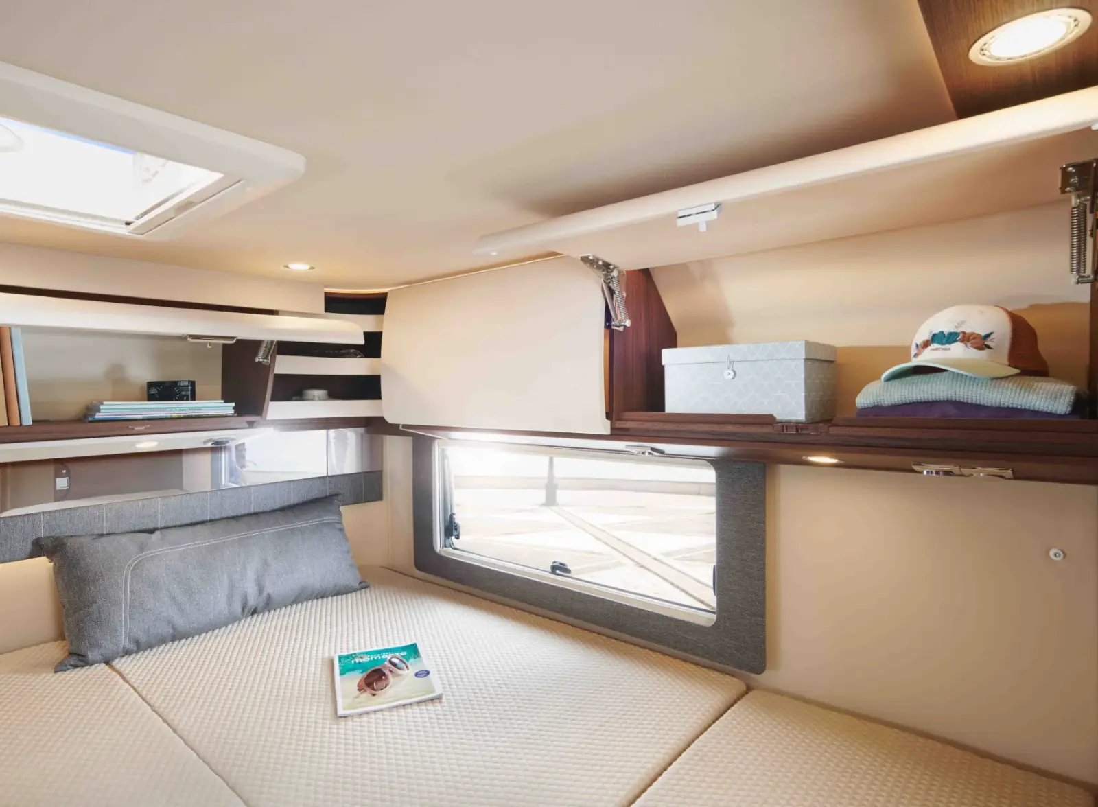 nnenansicht eines hellen und gemütlichen Wohnmobil-Schlafzimmers im Heck. Zu sehen ist ein großes Doppelbett mit cremefarbener, gesteppter Matratze, einem großen grauen Kissen und einer Zeitschrift mit Sonnenbrille darauf. Darüber befindet sich eine umlaufende Reihe von Oberschränken in edlem Holzdekor und cremefarbenen Fronten. Einige Klappen sind geöffnet und geben den Blick frei auf ordentlich verstaute Gegenstände wie eine graue Box, Kleidung und eine Basecap. Ein Seitenfenster und eine Dachluke lassen Tageslicht herein, ergänzt durch integrierte LED-Spotlights und Lichtleisten.