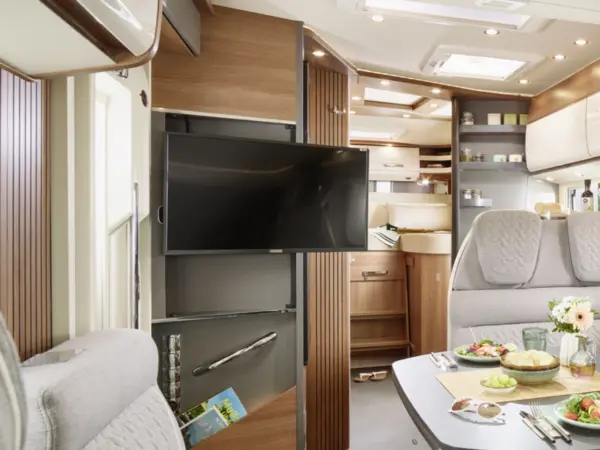 Modernes Wohnmobil-Interieur mit eingebautem Flachbildfernseher, Holzschränken und bequemen grauen Sitzmöbeln. Der Essbereich des Carthago C2-Tourer ist mit Tellern bestückt, während Küchengeräte und Regale das stilvolle Ambiente abrunden.