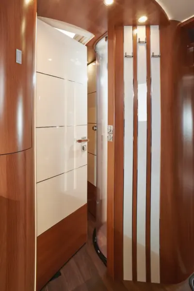 Ein modernes, kompaktes Badezimmer im Carthago Liner for Two zeichnet sich durch glänzend weiße und holzgetäfelte Wände, geschwungene Oberflächen, getäfelte Türen und eine runde Glasduschkabine aus, die von Deckeneinbauleuchten beleuchtet wird.