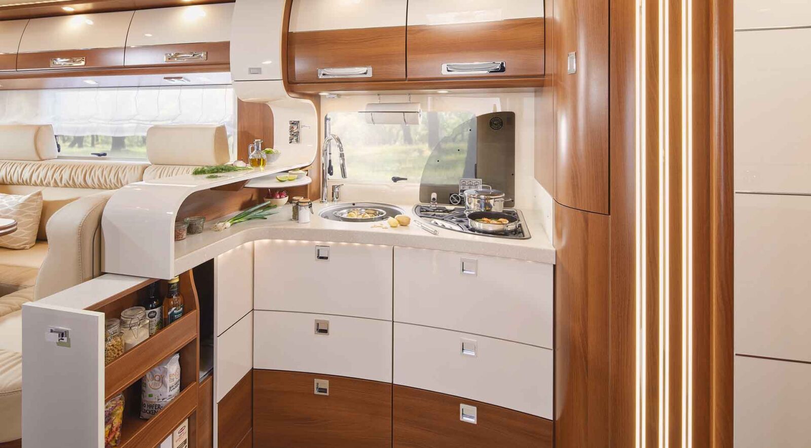 Moderne Wohnmobilküche in einem vollintegrierten Wohnmobil mit glänzend weißen Schränken, Holzakzenten, eingebautem Herd, Spüle, Unterschrankbeleuchtung, offenem Gewürzregal und einem Fenster mit Blick ins Grüne. Gemütliche Sitzecke ist teilweise sichtbar.