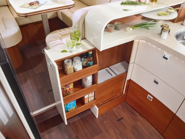 Die moderne Wohnmobilküche im Carthago Liner for Two ist mit Holzfußböden, cremefarbenen Arbeitsplatten und weißen Schränken ausgestattet. Eine ausziehbare Speisekammer bietet Platz für Gläser und Gewürze, während frische Kräuter und geschnittene Limetten den Tresen verschönern. Eine Sitzgruppe befindet sich im Hintergrund.