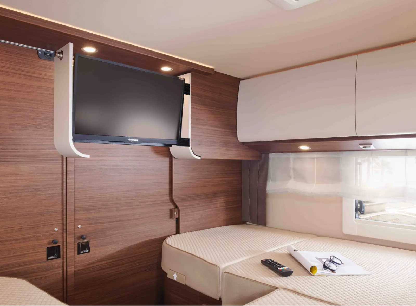 In einem holzgetäfelten Zimmer mit zwei Einzelbetten, das an das gemütliche Interieur eines teilintegrierten Wohnmobils erinnert, ist ein Flachbildfernseher angebracht. Auf einem Bett liegen Fernbedienung und Schlüssel, und durch das Fenster fällt natürliches Licht.