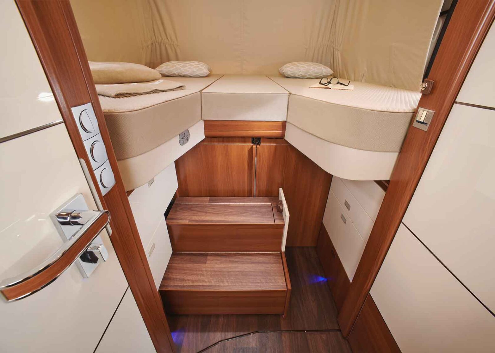 Kleines, gemütliches Schlafzimmer mit zwei Einzelbetten auf beiden Seiten, einer Holztreppe in der Mitte, Schubladen darunter und eine Brille und Kissen auf einem Bett, alles von einer offenen Tür aus gesehen.