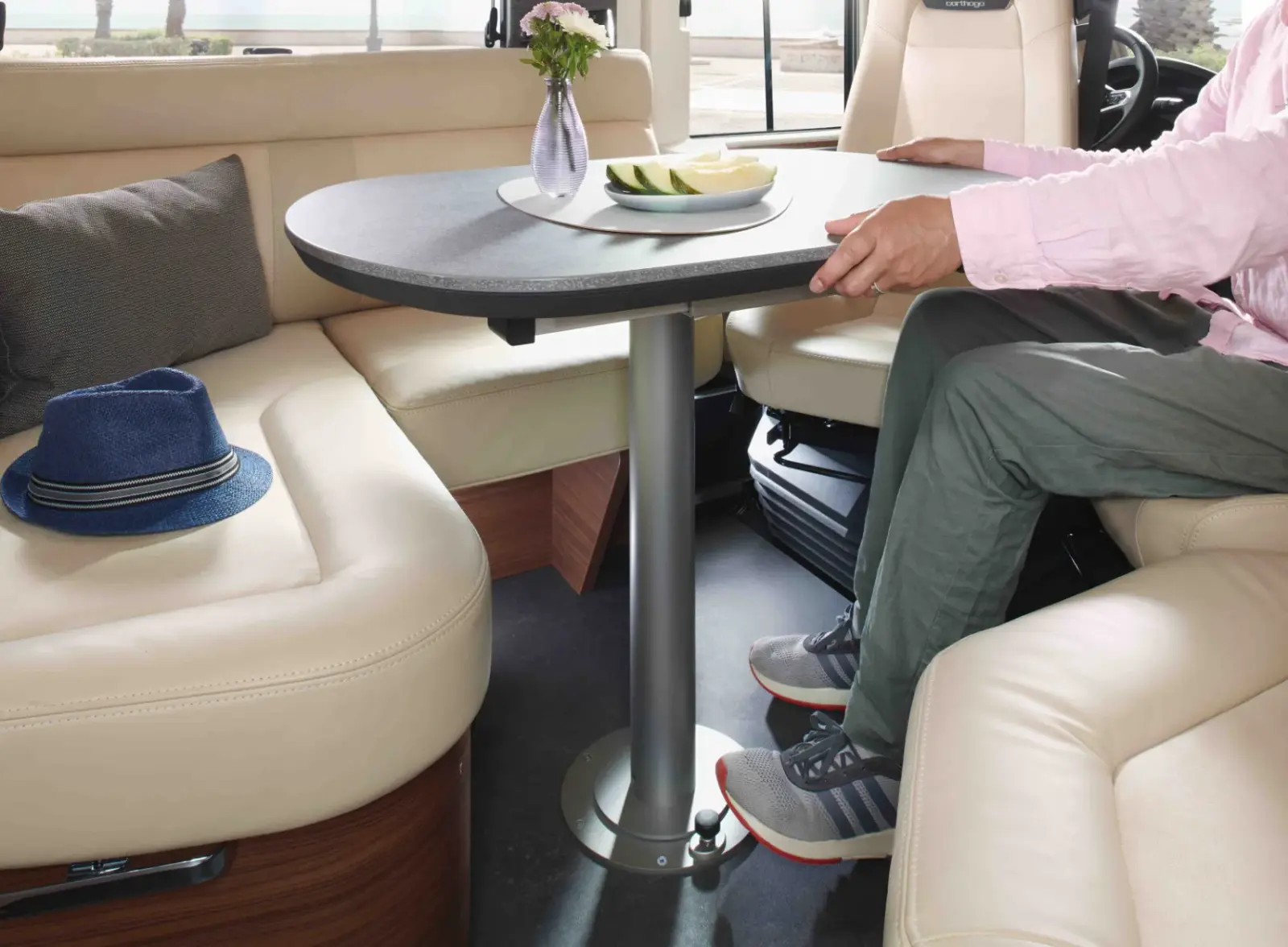 Eine Person sitzt an einem modernen, ovalen Tisch in einem Wohnmobil mit cremefarbenen Ledersitzen. Auf dem Tisch stehen ein Teller mit Melonenscheiben und eine kleine Vase mit Blumen. Ein blauer Hut liegt auf dem Sitz daneben.