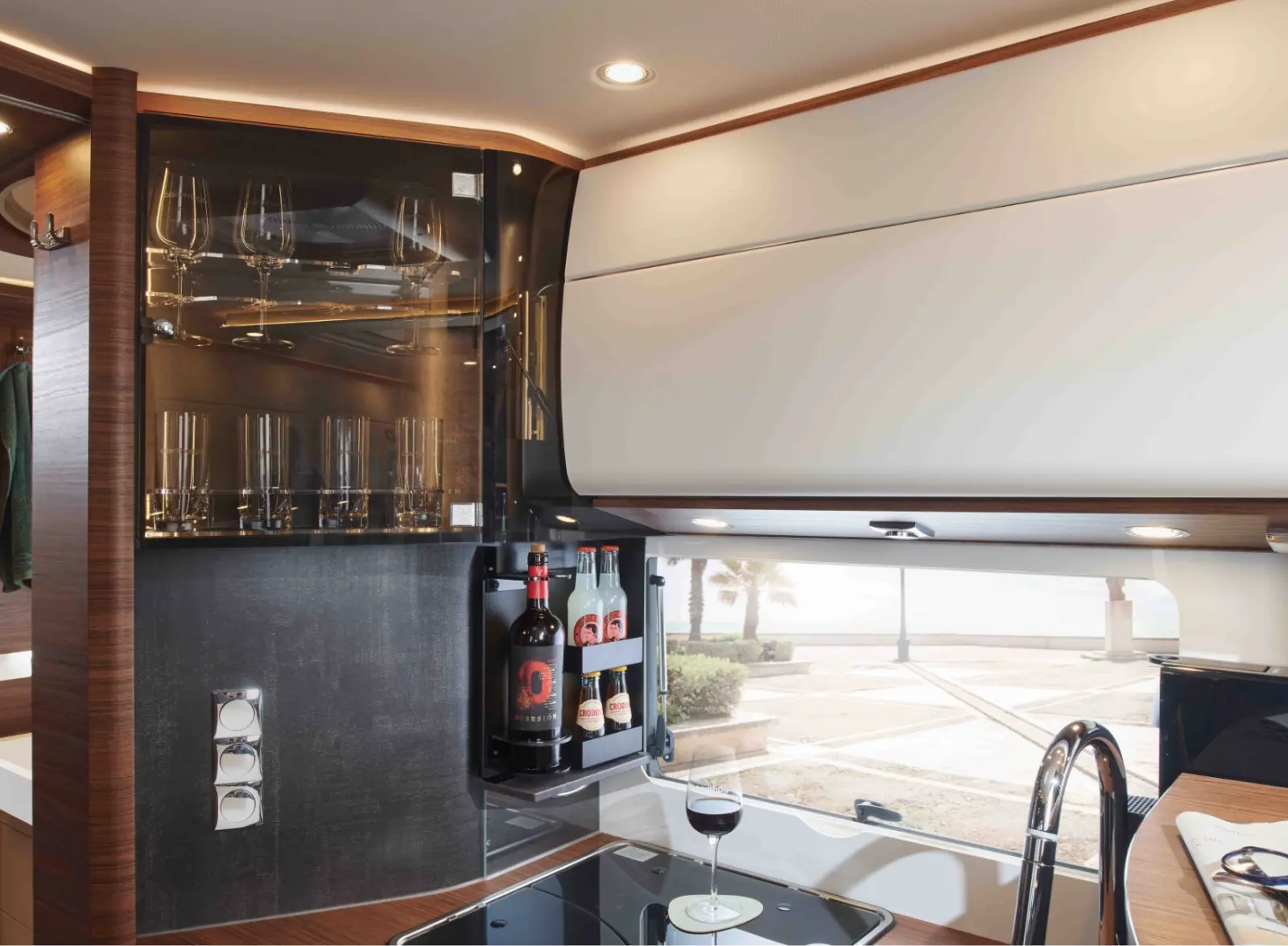 Eine moderne Wohnmobilküche in einem vollintegrierten Wohnmobil verfügt über eine Glasvitrine mit Weingläsern, ein Regal mit Flaschen und ein Glas Rotwein auf dem Tresen; ein großes Fenster gibt den Blick auf Bäume und Wasser frei.