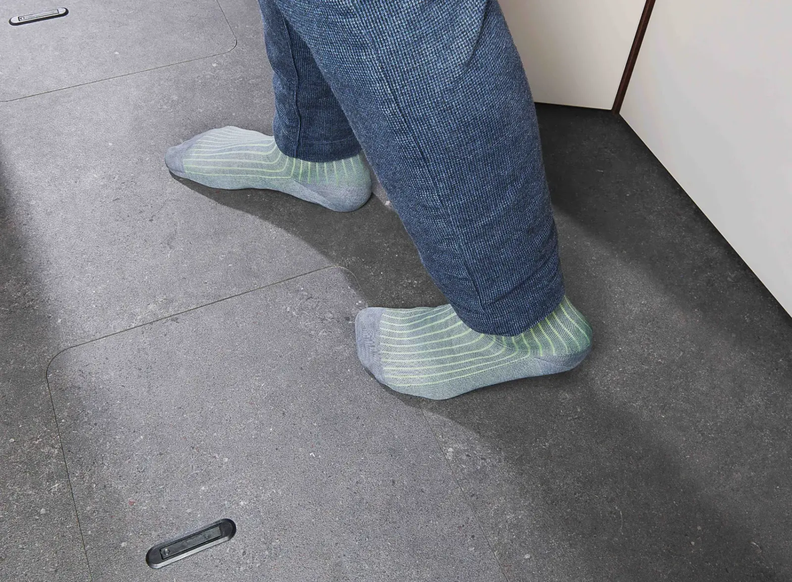Eine Person, die eine blaue Hose und hellgrün gestreifte Socken trägt, steht auf einem grauen Kachelboden neben einem hellen Schrank.