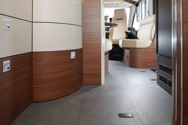 Modernes Wohnmobil-Interieur mit geschwungenen Holz- und cremefarbenen Stauschränken, grauem Bodenbelag und cremefarbenen Sitzen im vorderen Fahrbereich. Der Carthago Chic C Line T schafft einen sauberen, hellen und übersichtlichen Raum.