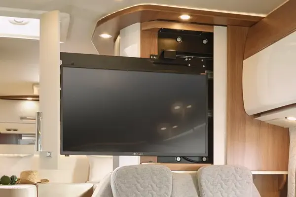 In einem modernen Carthago C Tourer Teilintegrierten Wohnmobil mit Holzakzenten, beigen Wänden und eingebauter Beleuchtung ist ein Flachbildfernseher auf einer drehbaren Halterung montiert. Im Vordergrund sind graue gepolsterte Sitze zu sehen.