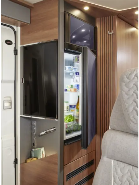Ein modernes Wohnmobil-Interieur eines Carthago C2-Tourer mit einem an der Wand montierten Fernseher, einem offenen Kühlschrank mit Lebensmitteln, Holzverkleidung und einem grauen gepolsterten Sitz im Vordergrund. Die helle Beleuchtung unterstreicht das schlichte, organisierte Design.