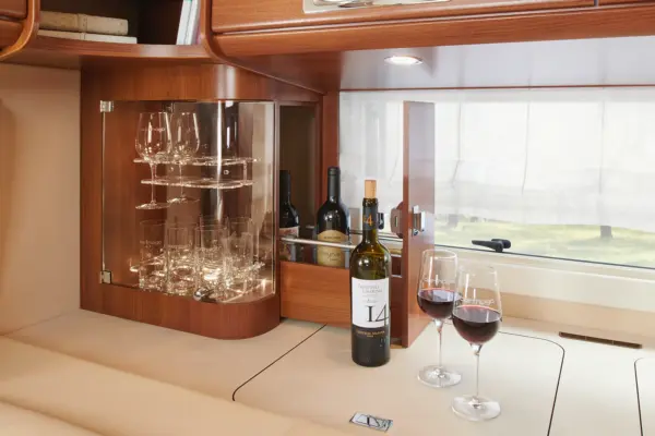 Ein Holzregal in einem gemütlichen Carthago Liner For Two zeigt Weingläser in einer Glasvitrine. Weinflaschen und zwei gefüllte Gläser stehen auf einer beigen Arbeitsplatte neben einem Fenster mit durchsichtigen Vorhängen.