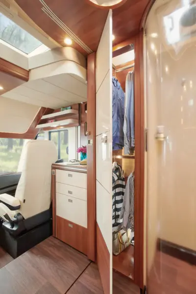 Modernes Wohnmobil-Interieur in einem Carthago Liner for Two, mit Holz- und Cremedekor, einem Kleiderschrank mit hängenden Kleidungsstücken, kompakten Küchenschubladen und dem Fahrersitz im Bug. Natürliches Licht strömt durch große Fenster und eine Dachluke.