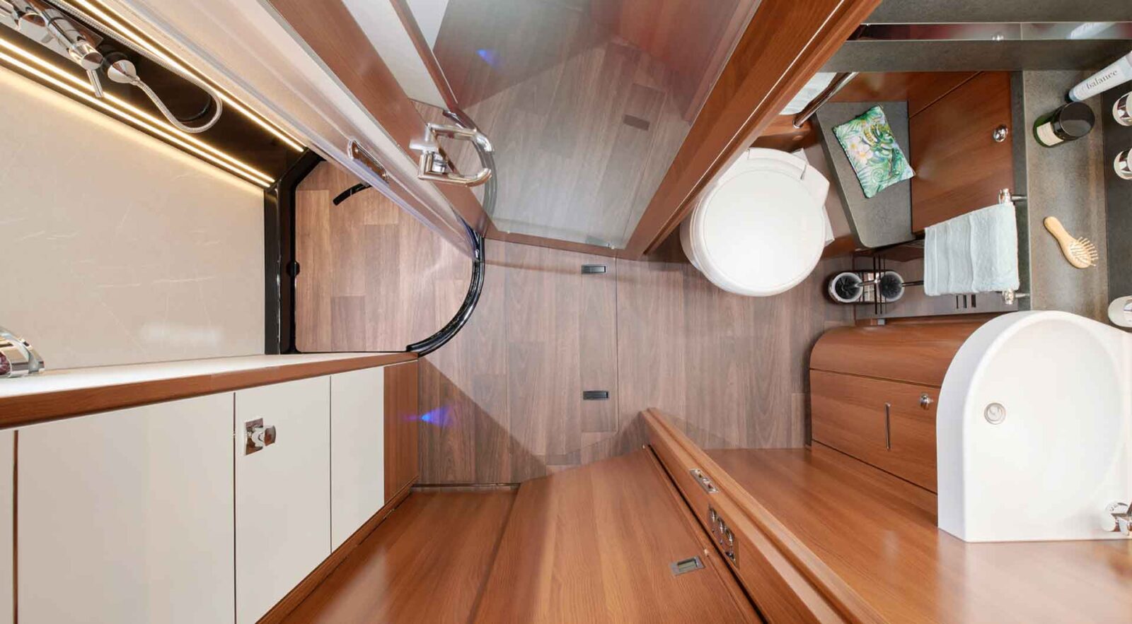 Ein modernes, kompaktes Badezimmer in einem vollintegrierten Wohnmobil mit Holzakzenten, einer weißen Toilette, einem Waschbecken, einer gebogenen Glastür in der Dusche und ordentlich angeordneten Toilettenartikeln und Handtüchern auf einer Arbeitsplatte. Das Foto ist von oben aufgenommen.