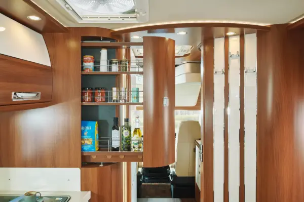Eine moderne Wohnmobilküche in einem Carthago Liner for Two mit Holzschränken und einer hohen ausziehbaren Speisekammer, die mit Konserven, Flaschen und Lebensmitteln gefüllt ist. Licht von oben beleuchtet den organisierten Stauraum.