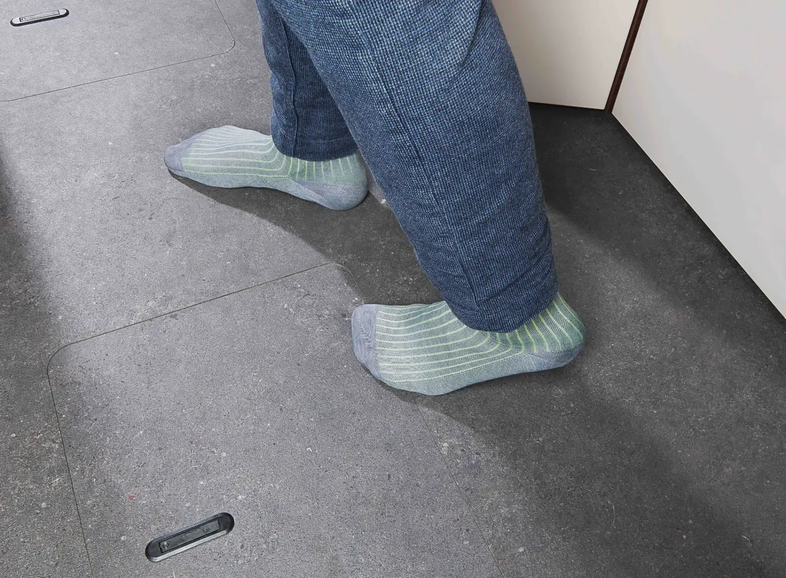 Eine Person mit blauer Hose und hellgrün gestreiften Socken steht auf einem dunkelgrauen Boden in der Nähe einer hellen Wand oder eines Schranks.