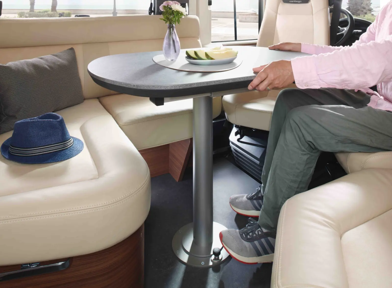 Eine Person sitzt an einem runden Tisch in einem modernen vollintegrierten Wohnmobil, mit beigen Ledersitzen, einem blauen Hut auf der Bank und einem Teller mit Obst und einer Blumenvase auf dem Tisch.