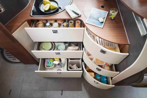 Eine moderne Carthago Chic C Line Küche mit mehreren offenen Schubladen und Schränken, in denen Geschirr, Gläser, Konserven, Snacks und Utensilien auf einer hölzernen Arbeitsplatte neben einem Waschbecken, einem Tablet, einer Zeitschrift und einem Glas mit einem Getränk angeordnet sind.