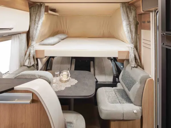 Der Innenraum eines Carthago C2-Tourer Wohnmobils besteht aus einem erhöhten Doppelbett mit Kissen, einem kleinen Esstisch mit zwei Gläsern und einer Kerze, gepolsterten Sitzbänken und gemusterten Vorhängen an den Fenstern.
