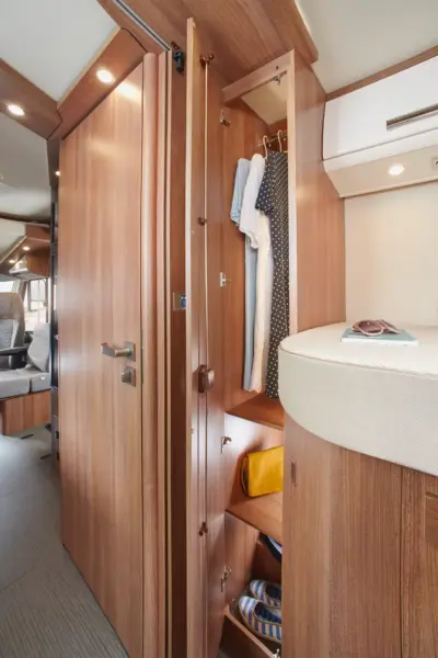Ein kompakter Kleiderschrank in einem Carthago C2-Tourer Wohnmobil mit Holzverkleidung, hängenden Kleidungsstücken, Regalen zur Aufbewahrung von Taschen und Zubehör sowie einem Teil des Bettes und des Wohnbereichs in der Nähe.