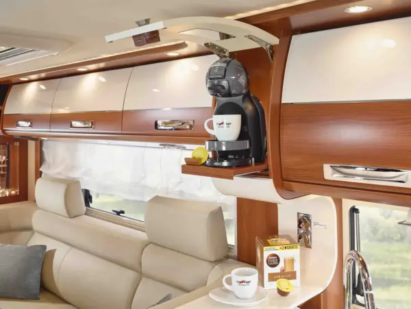 Ein modernes Carthago Liner for Two Wohnmobil-Interieur mit cremefarbenen Ledersitzen, einer Kaffeemaschine auf einem Holzregal, einer Tasse mit Untertasse, einer Schachtel mit Kaffeekapseln und einem Fenster mit weißen Vorhängen im Hintergrund.
