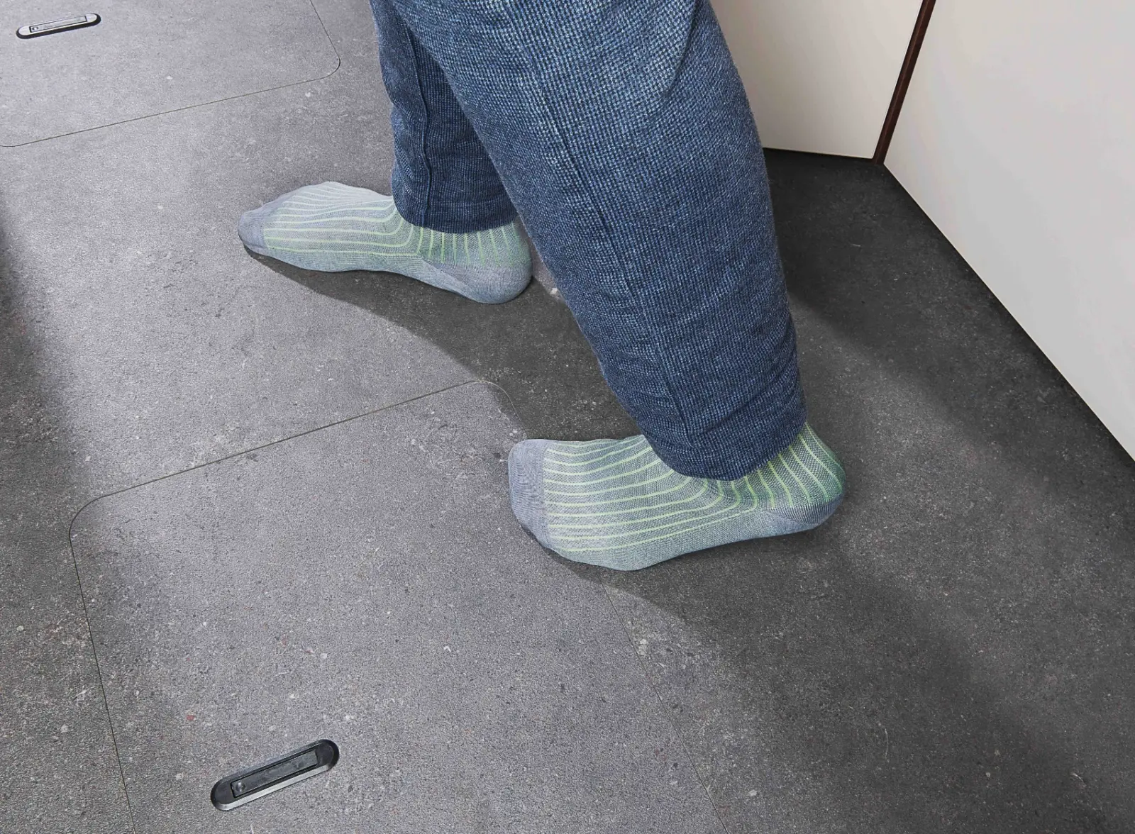 Eine Person mit hellgrün-weiß gestreiften Socken und blauer Hose steht auf einem grauen Kachelboden neben einem weißen Schrank in einem vollintegrierten Wohnmobil.