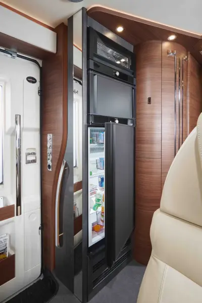 Ein modernes Wohnmobil-Interieur mit Holzvertäfelung, einem offenen Kühlschrank, der mit Lebensmitteln und Getränken gefüllt ist, einer eingebauten Mikrowelle darüber und einer sanften Beleuchtung, die eine gemütliche Atmosphäre schafft, verkörpert perfekt den Stil des Carthago Chic C Line T.