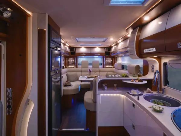 Modernes Wohnmobil-Interieur des Carthago Liner for Two mit eleganter Beleuchtung, weißen Schränken, Holzakzenten, einer geschwungenen Sitzecke mit Esstisch, einer Küchenzeile mit Spüle sowie verschiedenen Gerichten und Getränken auf dem Tresen und Tisch.