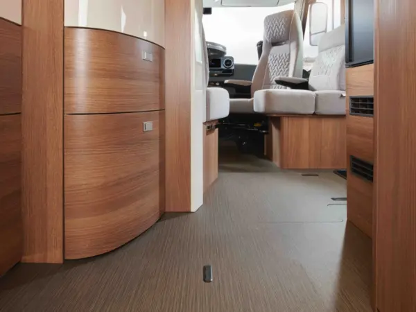Innenraum eines modernen Carthago C2-Tourer-Wohnmobils mit holzgetäfelten Schränken, einem strukturierten Boden, eingebautem Stauraum und zwei gepolsterten Fahrer- und Beifahrersitzen an der Vorderseite. Durch große Fenster fällt natürliches Licht ein.