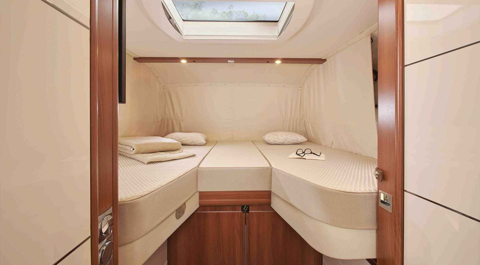 Ein kleines, ordentlich eingerichtetes Kabinenschlafzimmer in einem vollintegrierten Wohnmobil mit zwei Einzelbetten, Kissen, gefalteten Decken, eine Brille, Holzschränken unter den Betten und einem Oberlichtfenster darüber, das natürliches Licht hereinlässt.