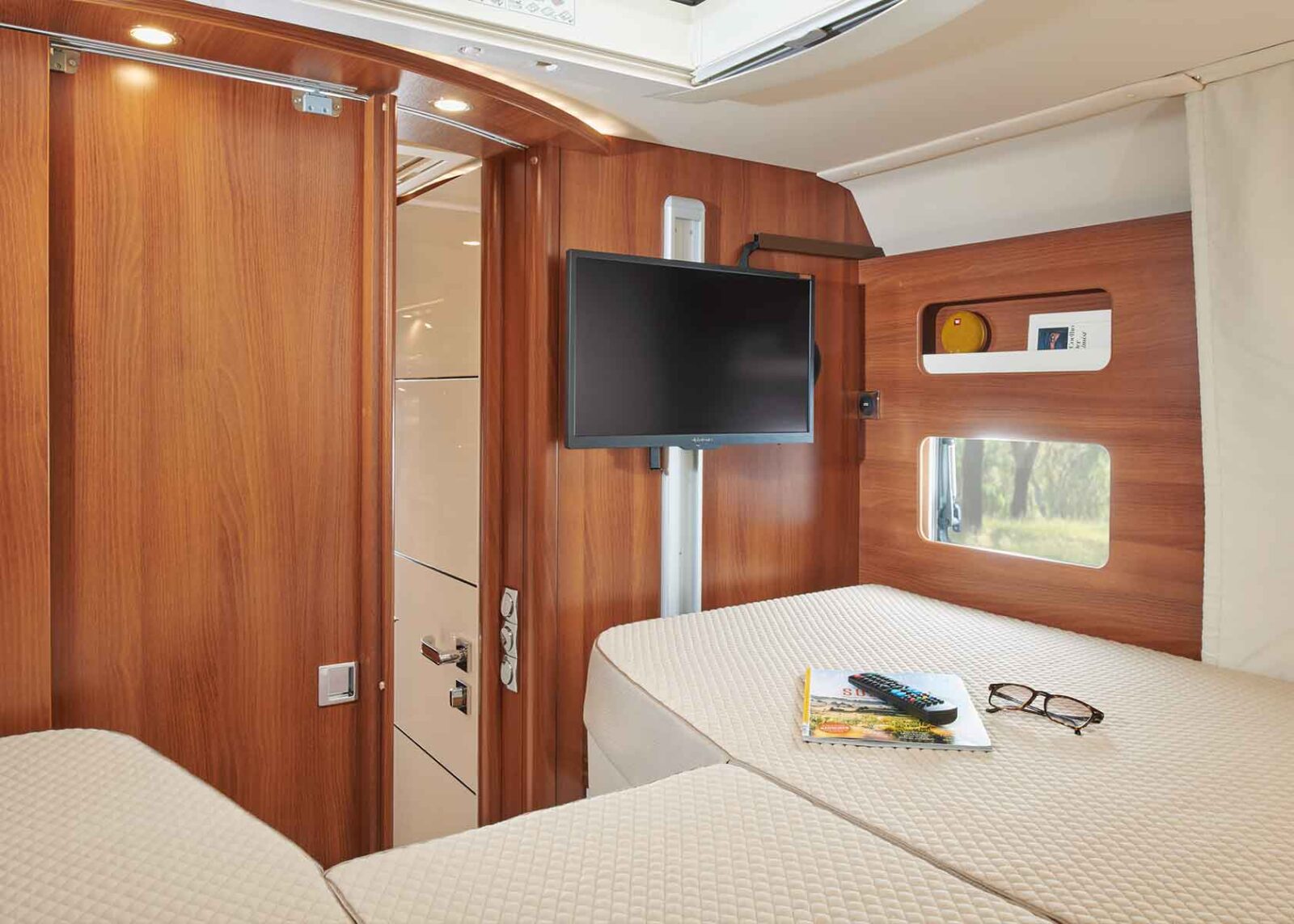 Innenraum eines modernen Wohnmobils mit Holzwänden, einem montierten Flachbildfernseher, zwei Einzelbetten und einem kleinen Regal mit Dekoration. Auf einem der Betten liegen eine Zeitschrift, eine TV-Fernbedienung und eine Brille.