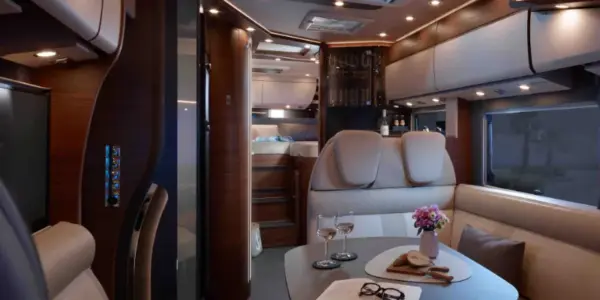 Luxuriöses teilintegriertes Wohnmobil-Interieur mit cremefarbenen Ledersitzen, einem gedeckten Esstisch mit Weingläsern, Blumen, Snacks und einem gemütlichen Bett im Hintergrund, alles warm beleuchtet von modernen Leuchten.