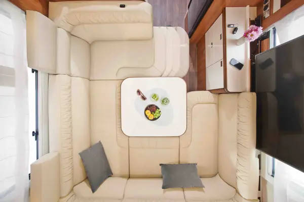 Draufsicht auf einen modernen Carthago Liner for Two Wohnmobil-Innenraum mit hellbeigen L-förmigen Sofas um einen quadratischen weißen Tisch mit Getränken und einem Teller mit Essen. Graue Kissen, ein Fernseher, Blumen und ein Telefon sind ebenfalls zu sehen.