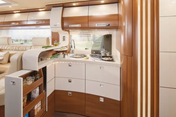 Moderne Wohnmobilküche mit hölzernen und weißen Schränken, einer Spüle, einem Herd mit Pfannen, Regalen mit Lebensmitteln und einem Essbereich im Hintergrund. Der gut beleuchtete und organisierte Raum unterstreicht die Eleganz des Carthago Liner for Two.