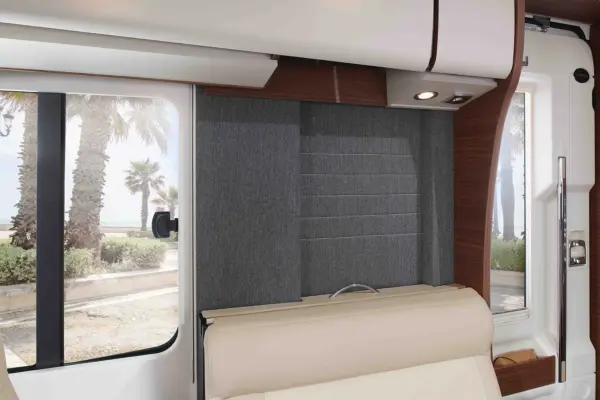 Innenraum eines modernen Carthago Chic C Line Wohnmobils mit beigen Sitzen, Holzakzenten und einem großen Fenster, das Palmen und einen Gehweg im Freien bei Tageslicht zeigt.