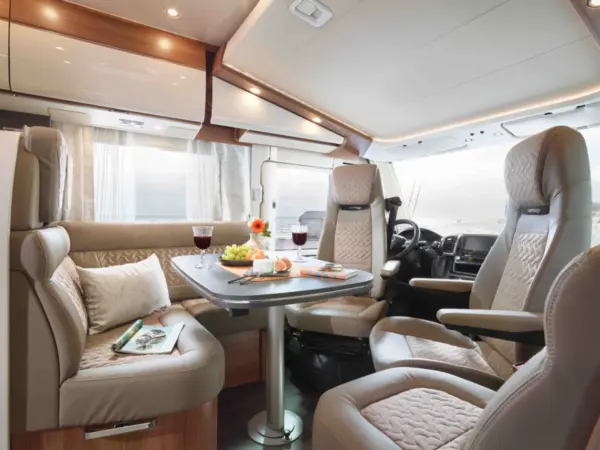 Modernes Wohnmobil-Interieur mit beigen Ledersitzen, einem Esstisch mit Snacks, Weingläsern, Blumen und einer Zeitschrift - dieser Carthago C2-Tourer bietet Komfort und Stil in der Nähe von großen Fenstern, die einen malerischen Blick nach draußen ermöglichen.