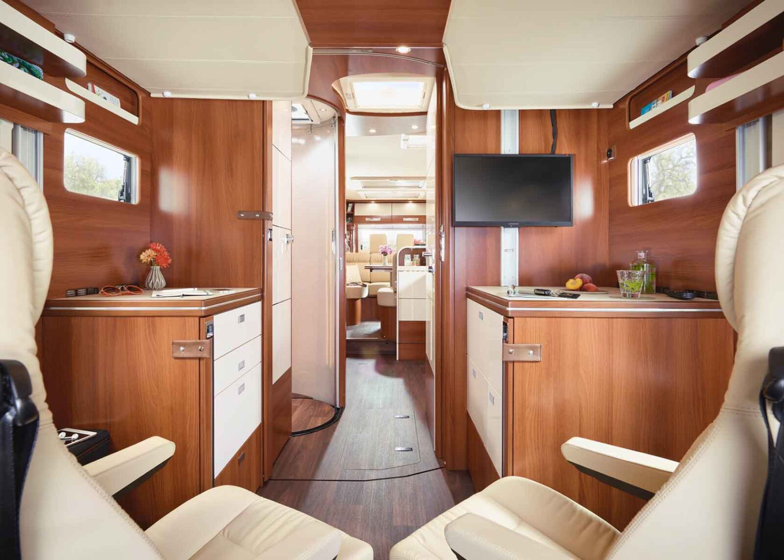 Innenraum eines modernen Wohnmobils mit Holzschränken, cremefarbenen Ledersitzen, einem an der Wand montierten Fernseher und einem kleinen Küchenbereich. Der Raum ist gut beleuchtet, organisiert und mit kleinen Pflanzen und Blumen dekoriert.