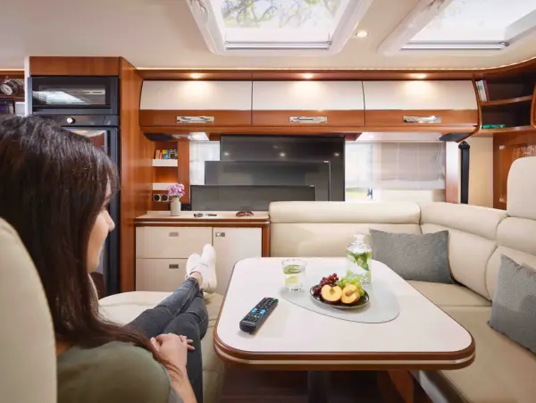 Eine Frau entspannt sich auf einem cremefarbenen Sofa in einem modernen Wohnmobil von Carthago Liner for Two vor einem Fernseher. Auf dem Tisch stehen Obst, Weintrauben, ein Glas mit Wasser, eine Fernbedienung und ein Glas. Die Einrichtung ist schlicht, mit Holzakzenten und Oberlichtern.