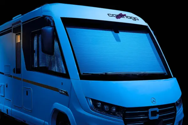 Ein modernes weißes Carthago C2-Tourer-Reisemobil mit einer großen Frontscheibe, die mit einer blau getönten Jalousie geschlossen und mit blauem Licht beleuchtet ist, wird vor einem dunklen Hintergrund gezeigt.