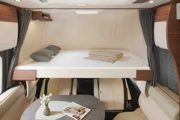 Der Innenraum eines modernen Carthago Chic C Line Wohnmobils besteht aus einem erhöhten Doppelbett mit Kissen und gefalteten Decken, cremefarbenem und holzfarbenem Dekor, einem kleinen Tisch mit einer Blumenvase und einer Zeitschrift auf dem Tisch.