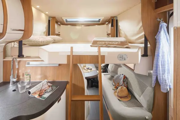Innenraum eines teilintegrierten Wohnmobils Carthago C Tourer mit erhöhtem Bett, Holzleiter, kompakter Küchenzeile und Sitzbereich mit Hut, Tasche und Hemd. Der effizient organisierte Raum ist für ein komfortables Leben auf Reisen gut beleuchtet.