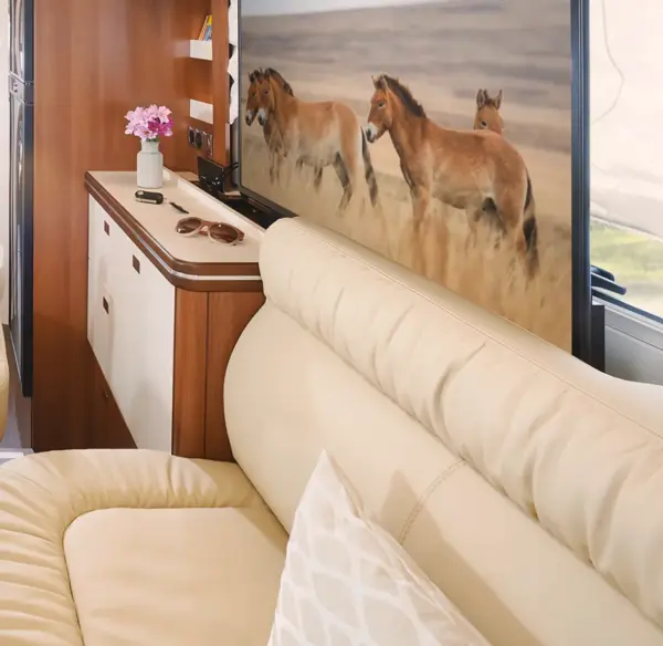 Ein gemütliches beigefarbenes Ledersofa in einem modernen Wohnmobil von Carthago Liner for Two steht vor einem Fernseher, auf dem Wildpferde laufen. In einem Schrank in der Nähe stehen rosafarbene Blumen, Sonnenbrillen und Dekoartikel, die einen komfortablen Lebensraum für die Reise schaffen.