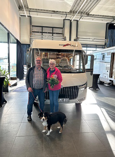 Ein lächelndes Paar steht vor einem weißen Wohnmobil beim Carthago Werksverkauf im Innenbereich. Die Frau hält einen Blumenstrauß in der Hand, während der Mann die Leine eines schwarz-weißen Hundes hält, der zu ihren Füßen sitzt. Das Sonnenlicht fällt durch große Fenster.