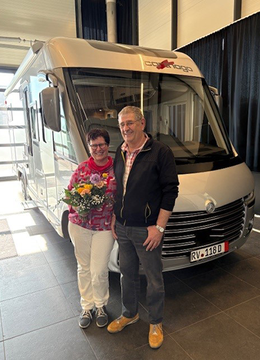 Ein lächelndes Paar steht im Innenbereich vor einem großen silbernen Wohnmobil im Carthago Werksverkauf. Die Frau hält einen Blumenstrauß in der Hand, und beide wirken glücklich und entspannt. Das Wohnmobil trägt ein Carthago-Logo und das Nummernschild RV-118 D.