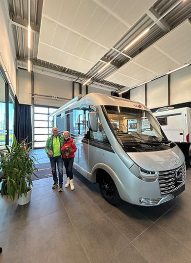 Zwei Menschen stehen lächelnd in einem modernen Ausstellungsraum neben einem großen silbernen Carthago Werksverkauf-Reisemobil. Die Umgebung ist hell mit hohen Decken, großen Fenstern und Pflanzen am Eingang.
