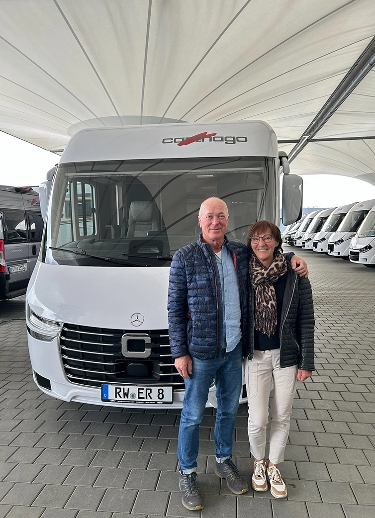 Ein älterer Mann und eine ältere Frau stehen lächelnd vor einem weißen Carthago Wohnmobil unter einem großen Vordach beim Carthago Werksverkauf und legen ihre Arme umeinander. Im Hintergrund sind mehrere andere Wohnmobile geparkt.