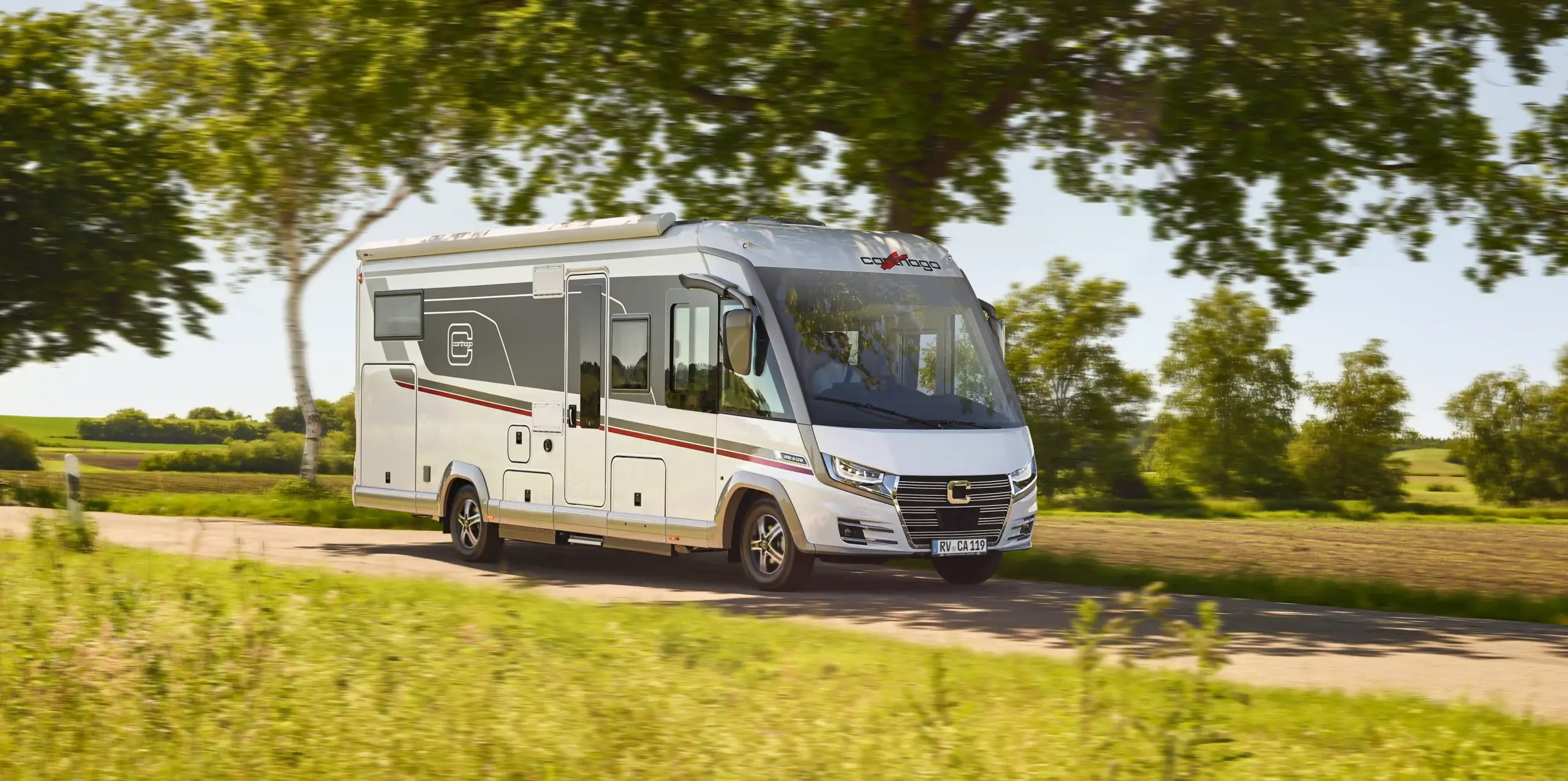 Ein modernes, silbernes Carthago Chic E Line Wohnmobil fährt an einem sonnigen Tag auf einer Landstraße entlang, die von grünem Gras und Bäumen gesäumt ist, mit klarem Himmel und Feldern im Hintergrund.